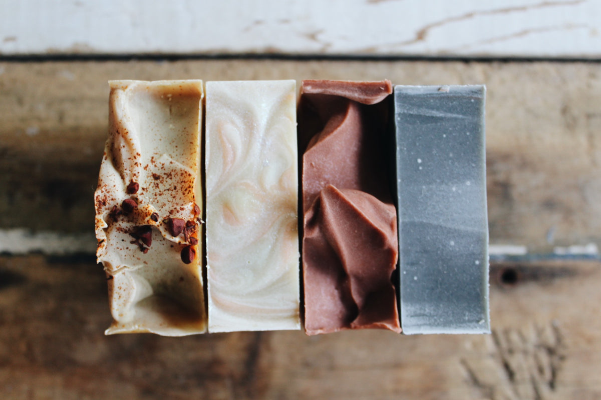Four artisanal handmade soap bars arranged vertically on a rustic wooden surface. The soaps feature distinct colors and textures: a light yellow bar with dark red specks, a cream, green and pink marbled bar, a dusty rose pink bar, and a speckled slate gray bar.