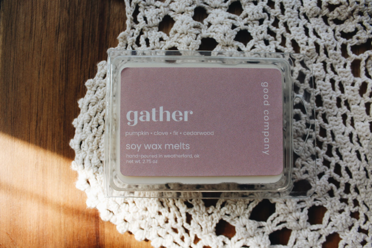 A clear plastic clamshell container of Good Company 'gather' pumpkin clove + cedarwood soy wax melts, resting on a white crocheted doily over a wooden surface.