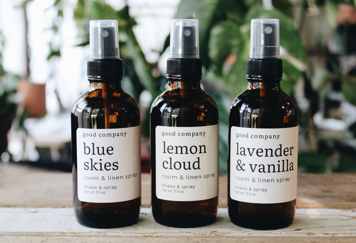 Three amber glass room and linen spray bottles with white labels on a rustic wooden surface, set against a blurred background of green houseplants. The labels read 'good company' and show three scents: 'blue skies', 'lemon cloud', and 'lavender & vanilla'.