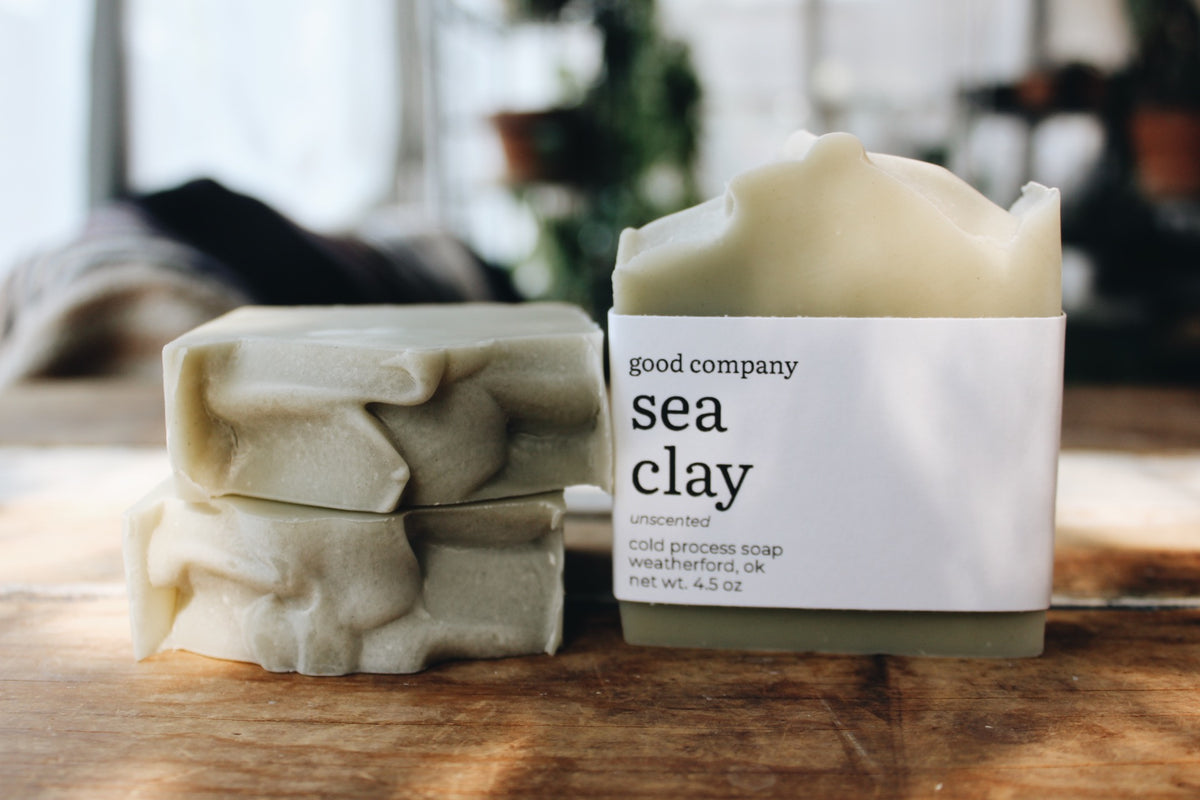 Two stacked bars of handmade soap in a muted sage green color and one in a paper sleeve label reading 'good company, sea clay, unscented, cold process soap, weatherford, ok, net wt. 4.5 oz', set on a wooden surface with a blurred indoor background.