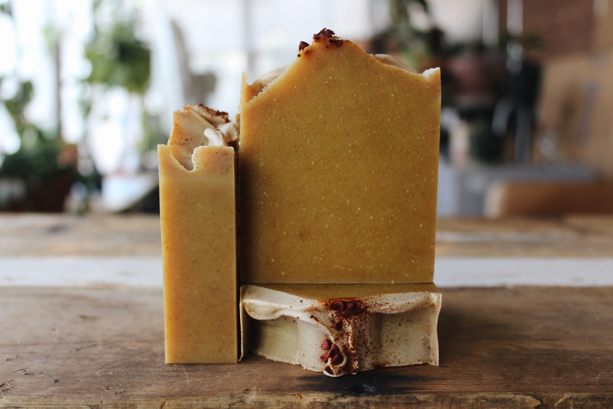 Three handmade bars of soap in warm golden-brown shades, standing upright on a rustic wooden surface with a softly blurred indoor background.