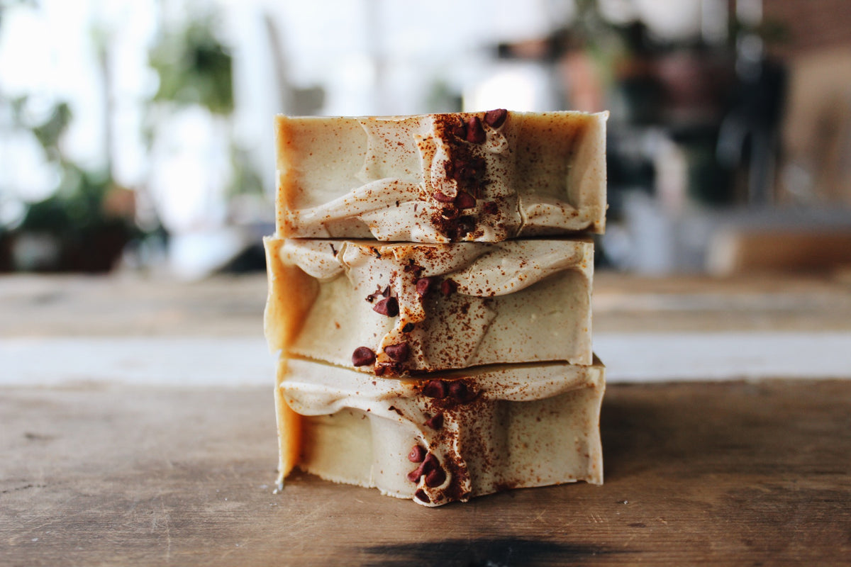 Three stacked bars of handmade soap with a creamy beige top and dried botanical inclusions, displayed on a rustic wooden surface with a soft-focus background.