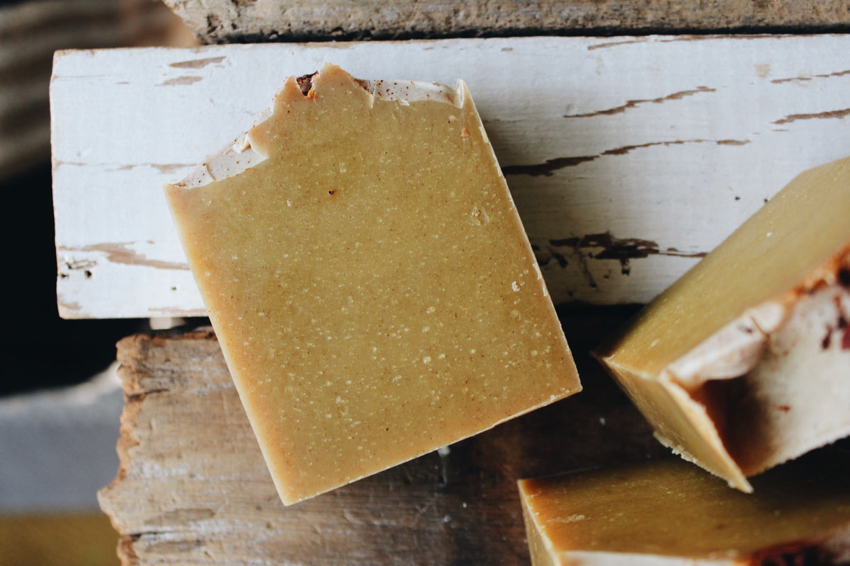 A close-up of a rustic handmade soap bar with a golden color, resting on weathered white and brown wooden planks.