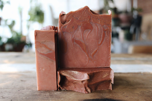 Three handmade soap bars in a dusty terracotta pink color with organic leaf-like swirls, arranged on a rustic wooden surface with a blurred background.