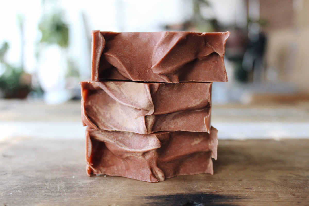 Three stacked rectangular bars of handmade soap in a warm reddish-brown color, on a wooden surface with a softly blurred indoor background.