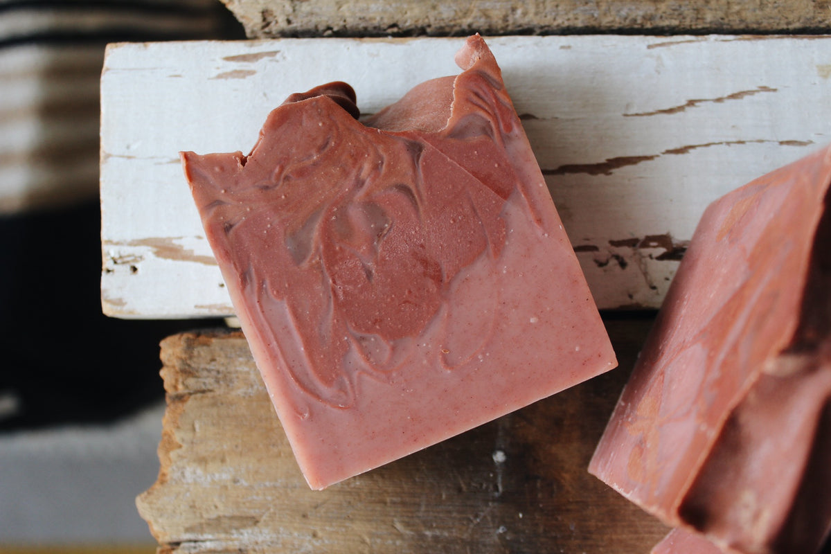 A close-up of a handmade rectangular bar of soap in a dusty rose pink color with marbled swirls, resting on a rustic white-painted wooden board.