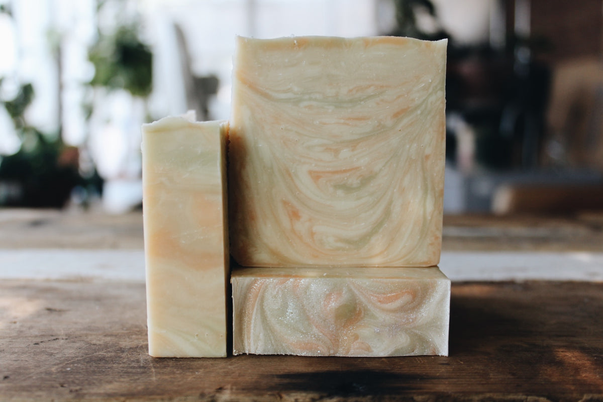 Three handmade soap bars with a marbled cream, green  and soft pink swirl pattern, arranged on a rustic wooden surface with a blurred indoor background.