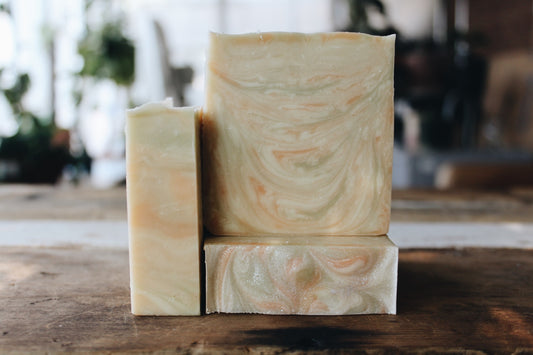 Three handmade soap bars with a marbled cream, green  and soft pink swirl pattern, arranged on a rustic wooden surface with a blurred indoor background.