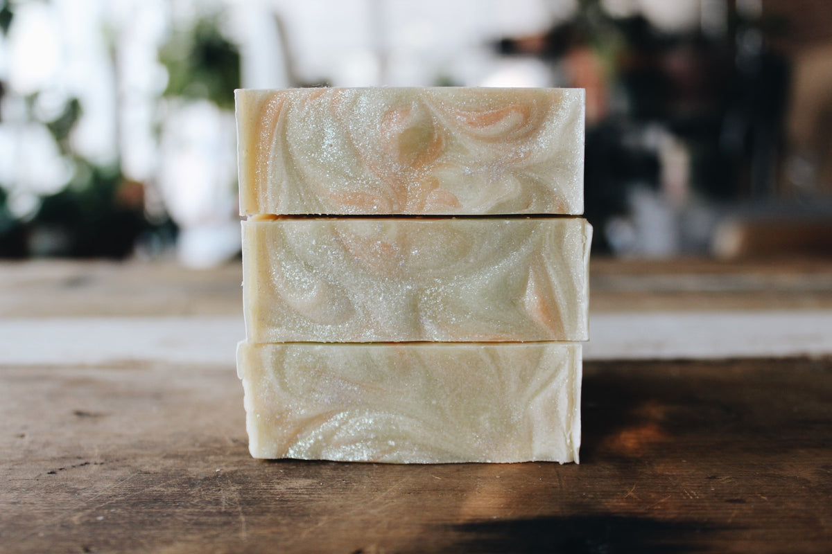 Three stacked rectangular bars of handmade soap with a creamy off-white base and soft peach swirls, standing on a rustic wooden surface with a blurred indoor background.