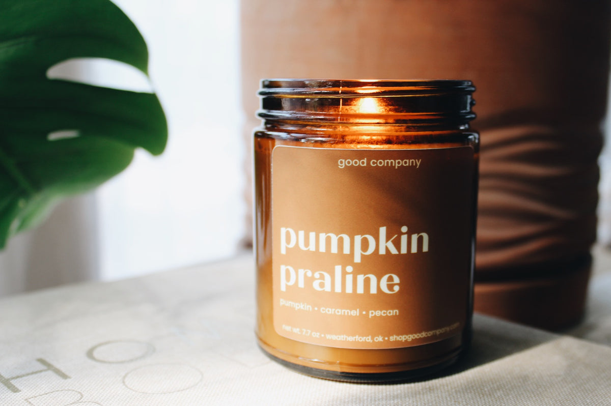 An amber glass jar candle labeled 'pumpkin praline' by Good Company, placed on a light surface with a blurred brown container and green leaf in the background.