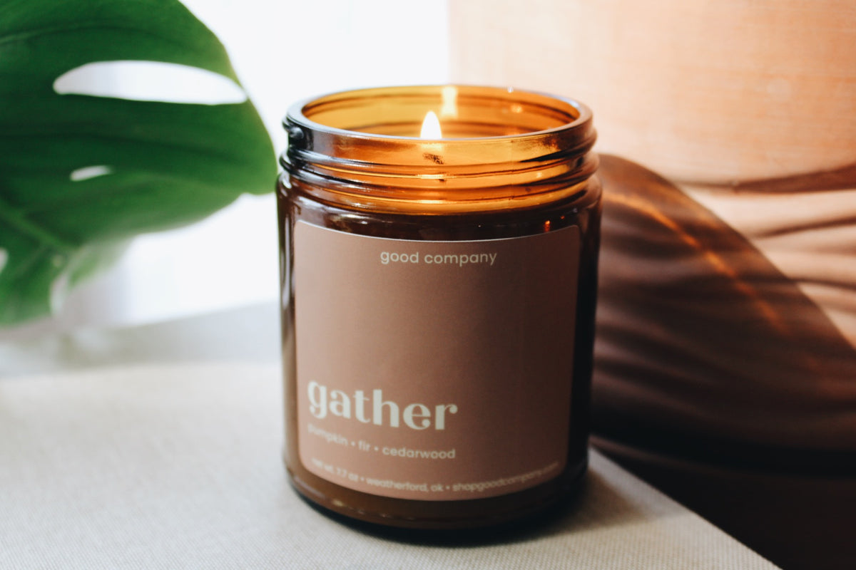 An amber glass jar candle with a lit flame and a taupe label reading 'gather' and 'good company', styled on a light surface with a green leaf and warm brown background elements.