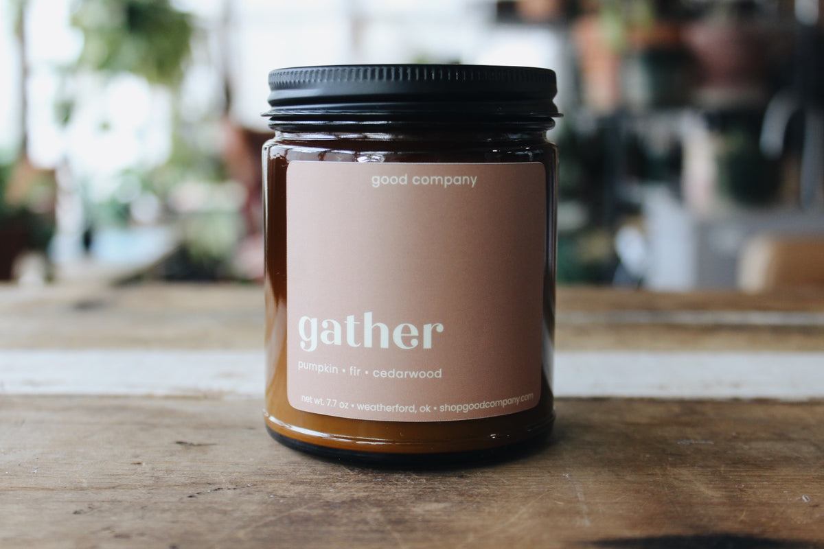 A glass jar candle with a black lid and a warm pink beige label reading 'gather' and 'good company', sitting on a rustic wooden table with a softly blurred indoor background.