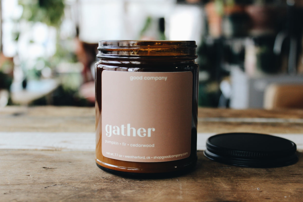 An amber glass jar candle with a pink beige label reading 'gather' and 'good company' at the top, sitting on a wooden table with its black lid placed beside it. The background is a softly blurred indoor setting.