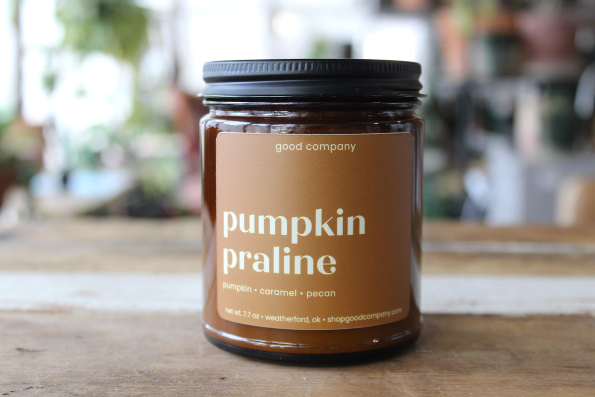 A glass jar candle with a black lid and a warm brown label reading 'pumpkin praline' and 'good company,' sitting on a wooden surface with a softly blurred indoor background.