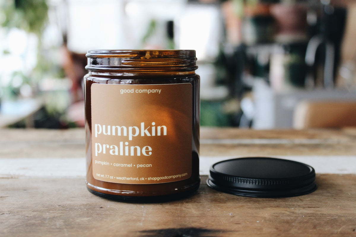 An amber glass jar candle labeled 'pumpkin praline' sits on a wooden table with its black lid placed beside it. The background is a softly blurred indoor setting with warm lighting.
