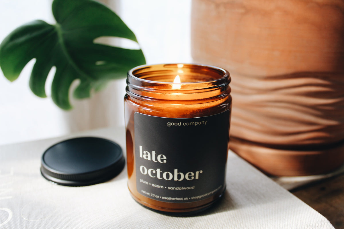 An amber glass jar with a lit candle with a plum label reading 'late october' and 'good company' is lit on a tabletop, with its black lid placed beside it. A green leaf and a large brown ceramic vase are softly blurred in the background.