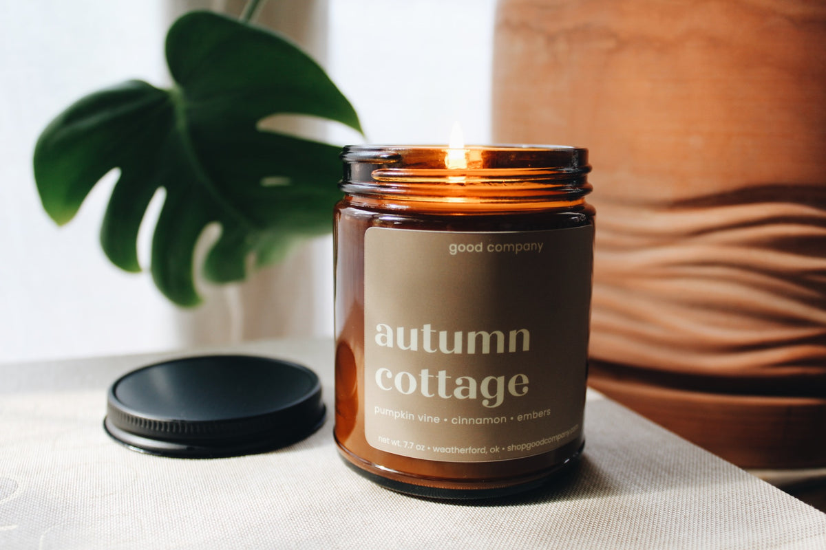An amber glass jar candle with a black lid resting beside it on a light fabric surface. The candle label reads 'good company' and 'autumn cottage,' with a lit flame visible inside.