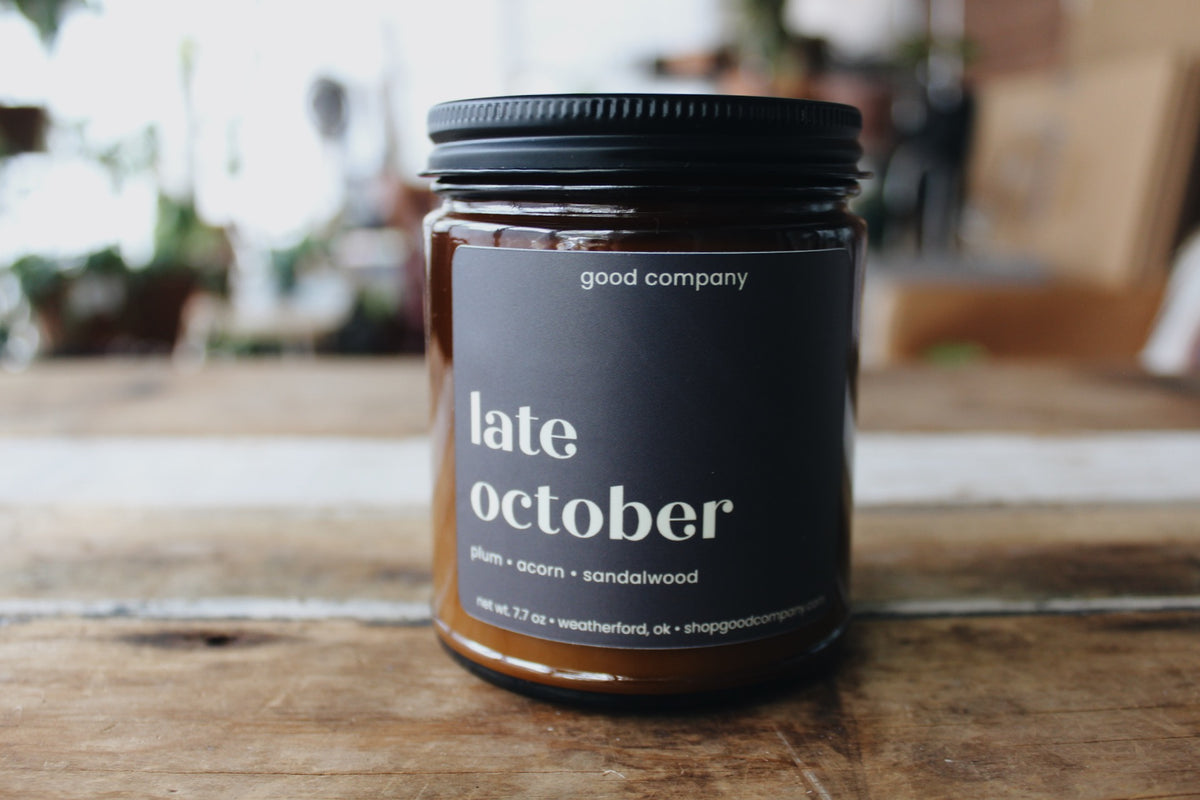 A glass jar candle with a black lid and a dark plum label reading 'late october' and 'good company,' sitting on a rustic wooden table with a softly blurred indoor background.