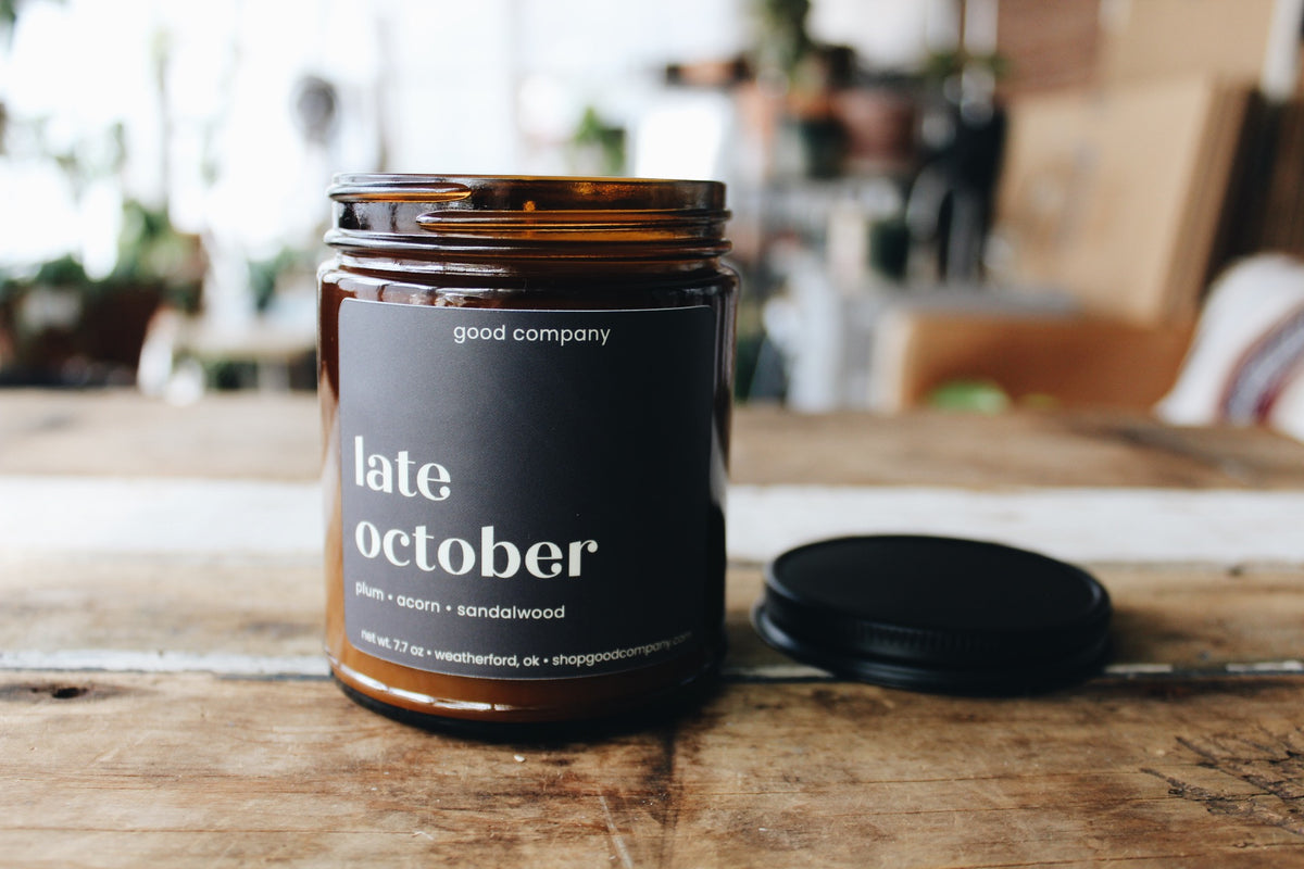 An amber glass candle jar with a dark plum label reading 'late october' and 'good company' at the top, shown on a rustic wooden table with its black lid placed beside it. The background is softly blurred, showing an indoor setting.