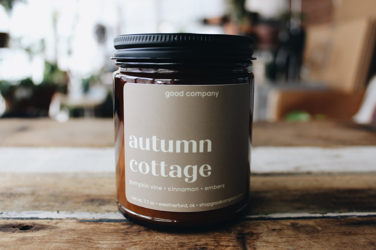A glass jar candle with a black lid and a grey label reading 'autumn cottage' and 'good company', placed on a rustic wooden surface with a softly blurred indoor background.