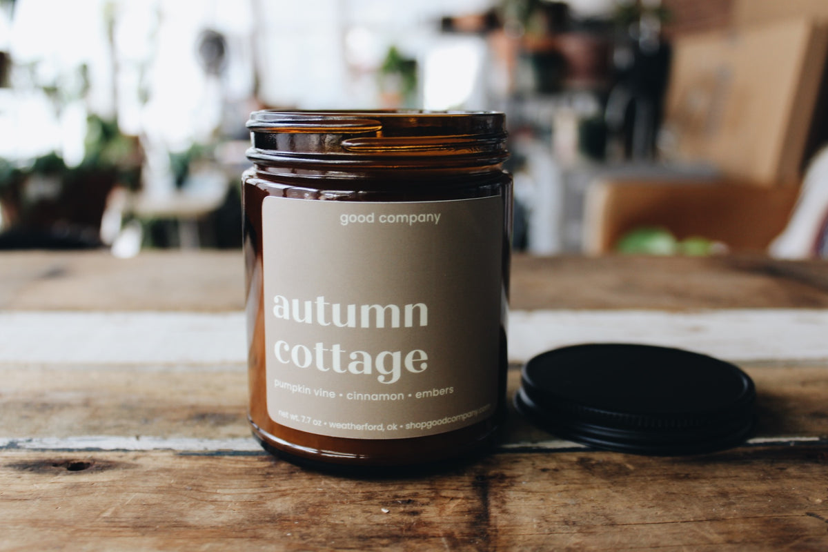 An amber glass jar candle labeled 'autumn cottage' by good company, sitting on a rustic wooden table with its black lid placed beside it. The background is a softly blurred indoor setting.
