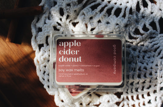 A clear plastic clamshell package of Good Company soy wax melts labeled 'apple cider donut' in a dusty rose color, placed on a white crocheted textile over a wooden surface.
