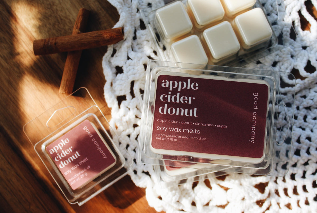 Good Company soy wax melts in the flavor apple cider donut, shown in clear plastic clamshell packaging with a dark burgundy label. One pack is open, showing the wax melts inside, and another pack sits nearby.