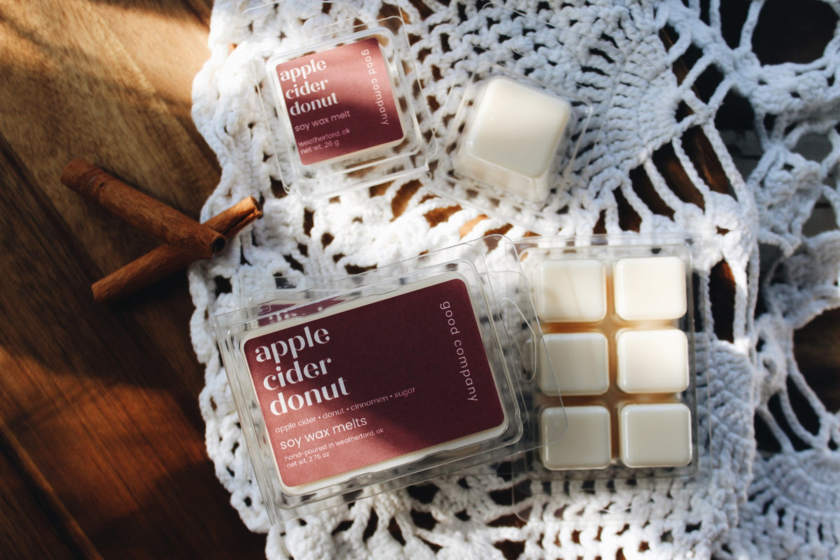 Two clear plastic containers of apple cider donut scented soy wax melts are arranged on a white crocheted lace cloth over a wooden surface, with cinnamon sticks nearby. One container is closed with a dark burgundy label, and the other is open, showing white wax cubes.