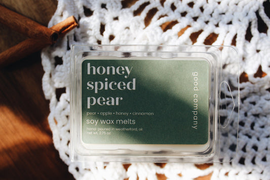 A clear plastic clamshell package of Good Company soy wax melts labeled 'honey spiced pear' sits on a white crocheted textile over a wooden surface with cinnamon sticks.