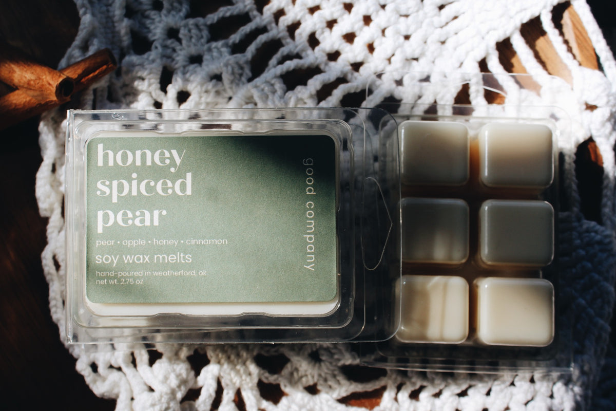 A package of Good Company soy wax melts labeled 'honey spiced pear' shown next to six individual wax cubes in a clear plastic clamshell container, styled on a white crocheted textile with warm brown background elements.