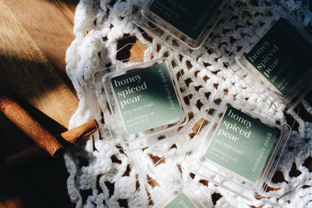 Several clear plastic clamshell packages of Good Company 'honey spiced pear' soy wax melts are arranged on a white crocheted doily over a wooden surface, with warm sunlight casting shadows.