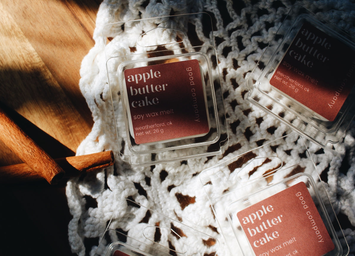 Several clear clamshell wax melts labeled 'apple butter cake' are arranged on a white crocheted textile over a warm wooden surface. The labels are dusty rose pink with white text, and the scene is lit with strong natural sunlight and shadows.