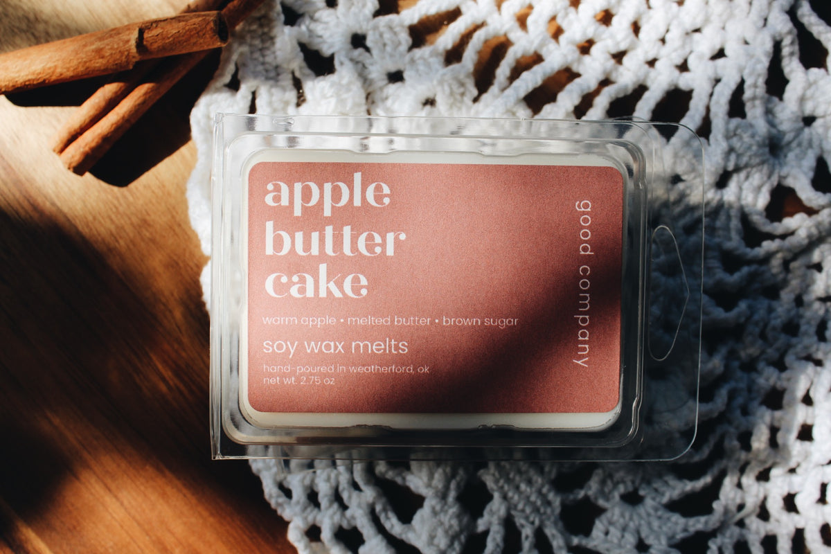 A packaged soy wax melt labeled 'apple butter cake' by Good Company, shown in a clear plastic clamshell with a dusty rose label. The package is photographed on a white crocheted textile over a wooden surface with cinnamon sticks, creating a styled flat-lay scene.