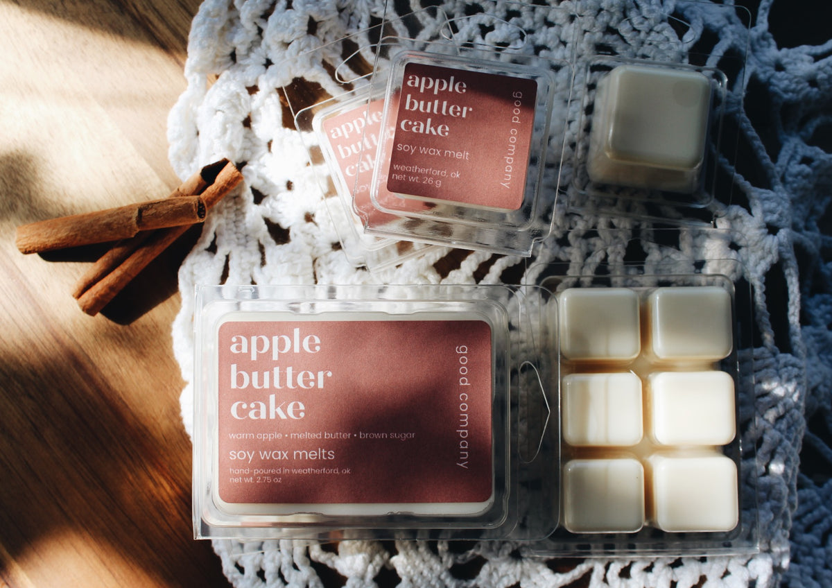 Two packages of Good Company 'apple butter cake' soy wax melts are arranged on a white crocheted cloth with cinnamon sticks on a wooden surface. One package shows the pinkish-brown wax melt labels, and the other shows several off-white wax melts inside clear plastic packaging.