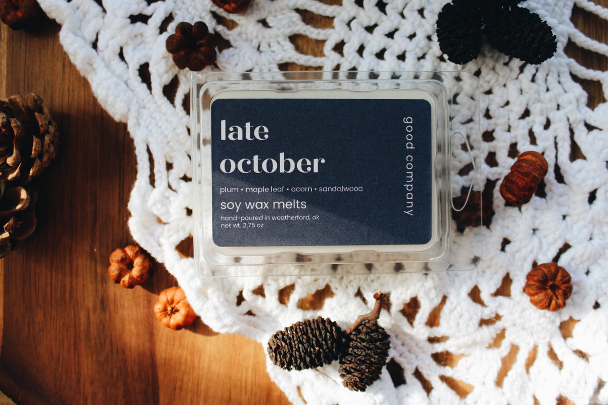 A clear plastic clamshell container of Good Company 'late october' soy wax melts sits on a white crocheted doily over a wooden surface, surrounded by pinecones and small orange pumpkins.