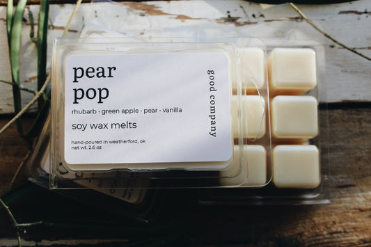A clear plastic clamshell package of Good Company 'pear pop' soy wax melts, showing 6 pale cream-colored wax cubes and a white label with black text, set on a rustic wooden surface.