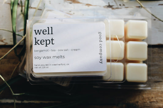 A clear plastic clamshell package of Well Kept soy wax melts with a white label, showing 6 cream-colored wax cubes inside. The package is placed on a rustic wooden surface with dried greenery in the background.