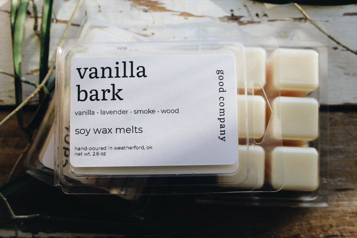 A clear plastic clamshell of Good Company Vanilla Bark soy wax melts, showing 6 cream-colored soy wax cubes inside. The front features a white label with black text that reads, 'vanilla bark, vanilla, lavender, smoke, wood, soy wax melts, hand-poured in weatherford, ok, net wt. 2.75 oz' and the package is styled on a rustic wooden surface with a plant in the background.