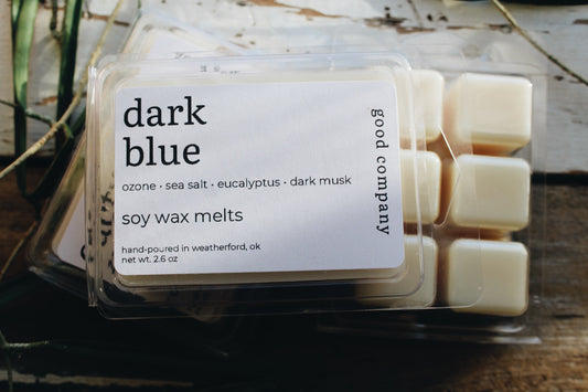 A package of Good Company 'dark blue' soy wax melts, showing the cream-colored wax cubes inside a clear plastic clamshell container with a white label, set on a rustic wooden surface.