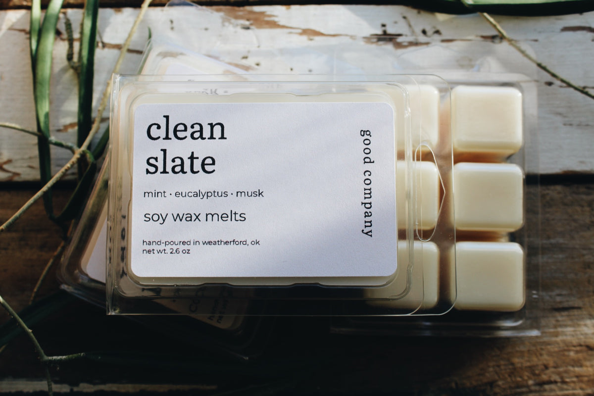 A clear plastic clamshell package of Good Company Clean Slate soy wax melts, showing the cream-colored wax cubes inside, set on a rustic wooden surface with dried greenery.