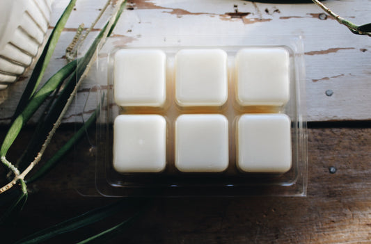 Six square cream wax melts in a clear plastic clamshell package, arranged in a 2x3 grid on a rustic white and dark wood surface with green leaves.