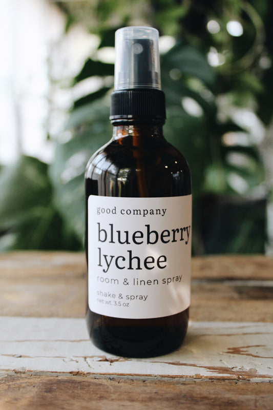 A 3.5 oz amber glass spray bottle of 'good company' Blueberry Lychee Room & Linen Spray, featuring a white label and black pump, set on a rustic wooden surface with a blurred green plant background.
