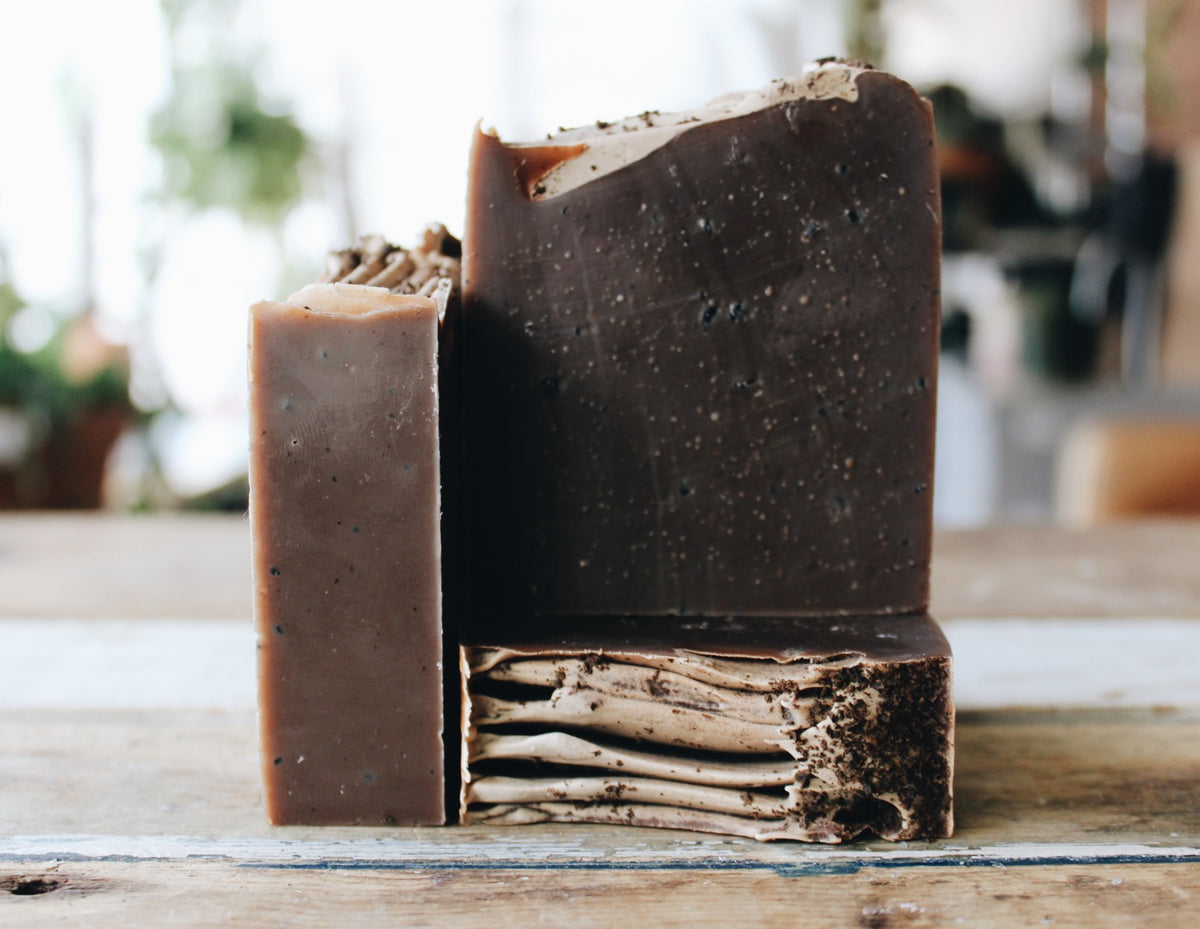 Three handmade soap bars in earthy brown and dark brown shades, displayed on a rustic wooden surface with a softly blurred indoor background.