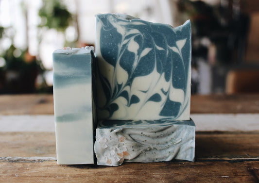 Three handmade marbled soap bars in blue, white, and gray tones, stacked on a rustic wooden surface with a softly blurred indoor background.