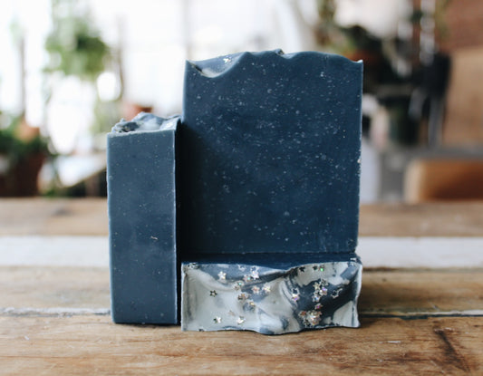 Three handmade rectangular bars of soap in a dark slate blue color with silver biodegradable star glitter on top, standing on a rustic wooden surface with a blurred indoor background.