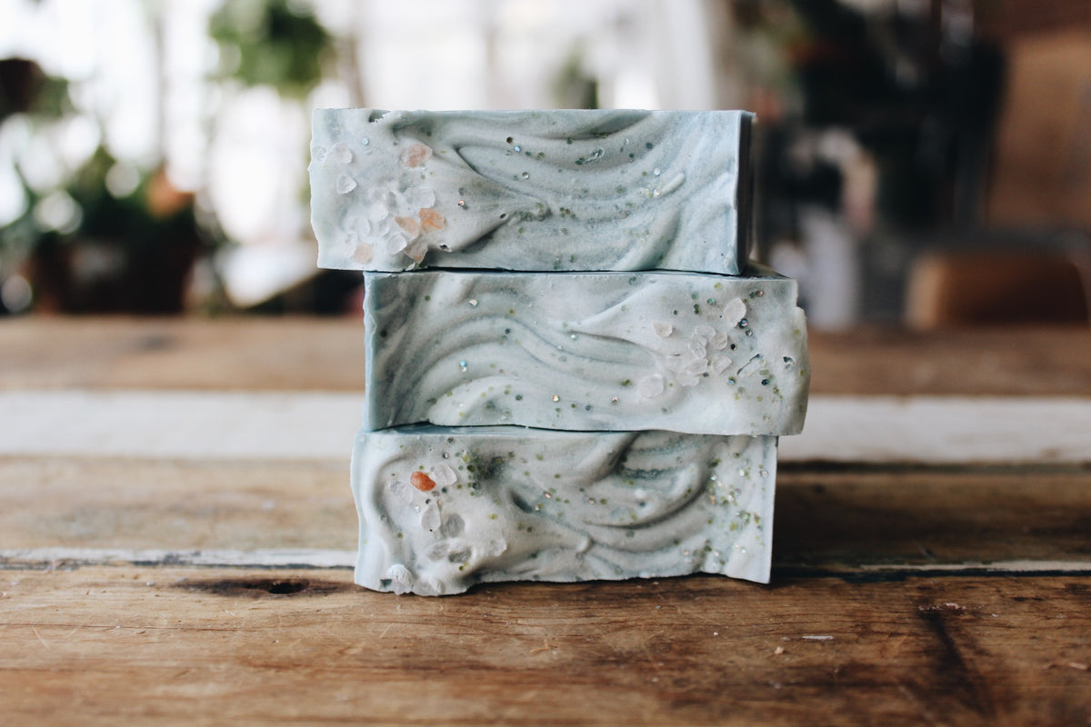 Three stacked handmade soap bars with a swirled light blue-gray pattern and embedded botanical specks, resting on a rustic wooden surface.