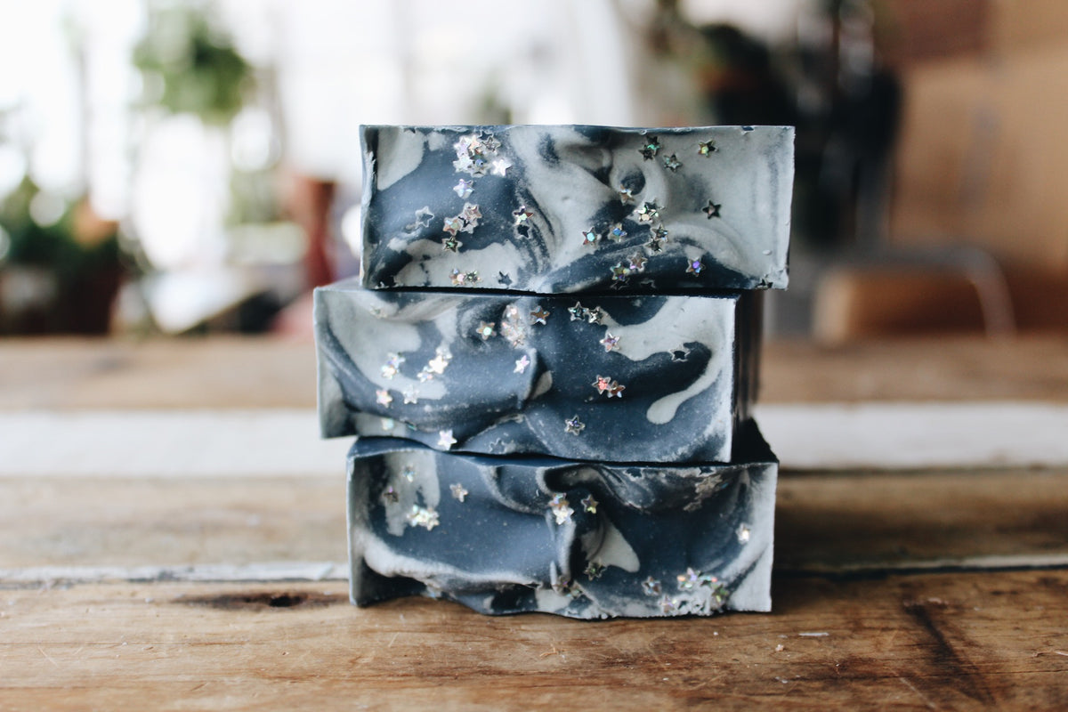 Three stacked rectangular bars of soap with a dark gray and white marbled pattern and small silver star inclusions, displayed on a rustic wooden surface with a blurred indoor background.