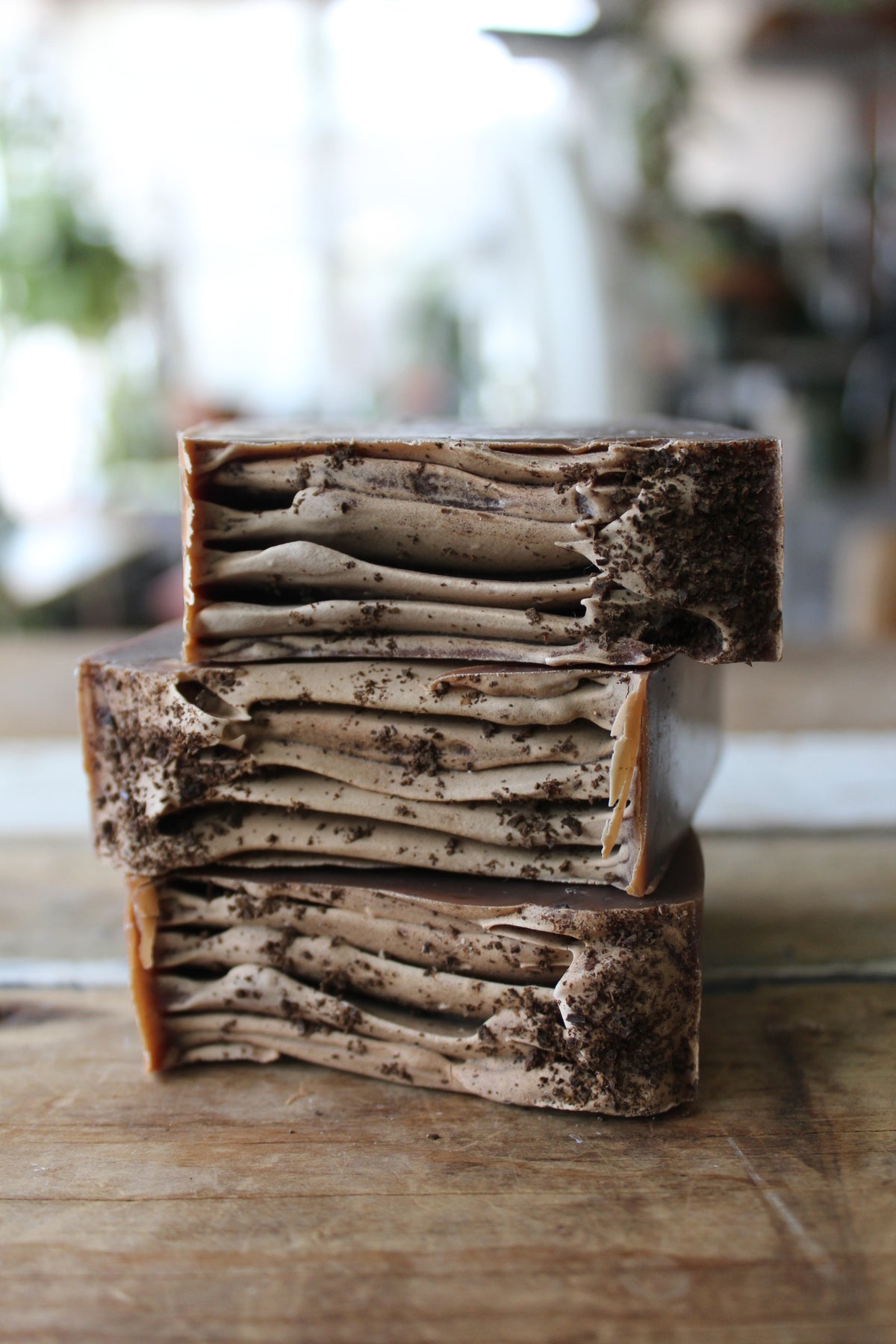 Three stacked rectangular bars of handmade soap with a layered pattern of beige and dark brown, standing on a rustic wooden surface with a blurred indoor background.
