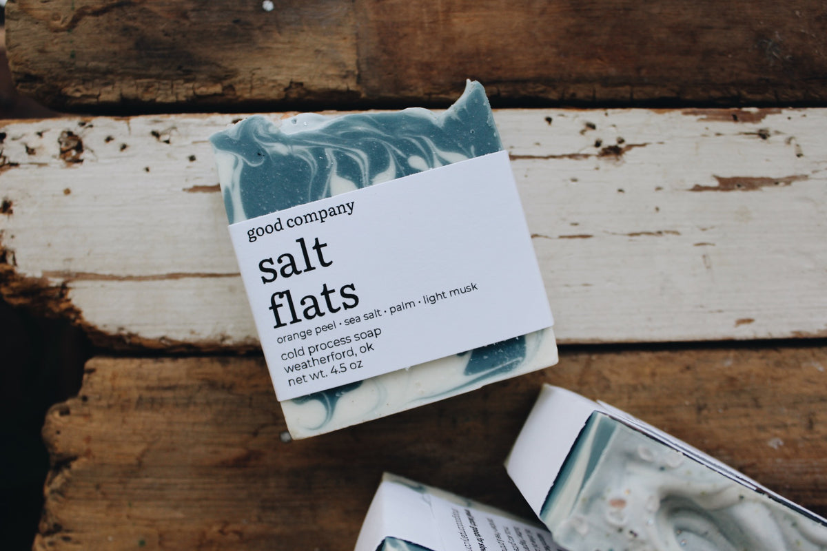 A bar of 'salt flats' cold process soap by 'good company' resting on a rustic, weathered wooden surface. The soap has a marbled blue and white swirl pattern, wrapped in a white paper label with black text.