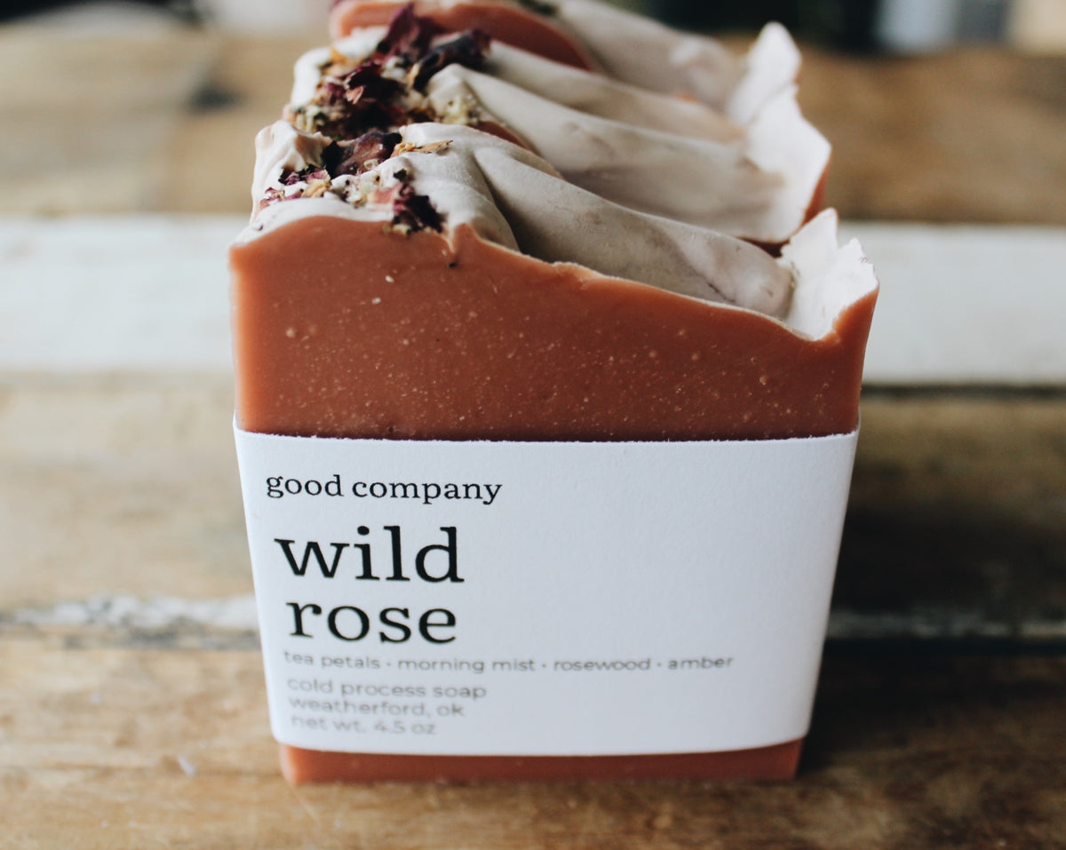 A bar of handmade soap labeled 'good company wild rose' with a white paper wrapper, showing a rose-colored base and layered cream-colored top with dried flower petals, set on a rustic wooden surface.